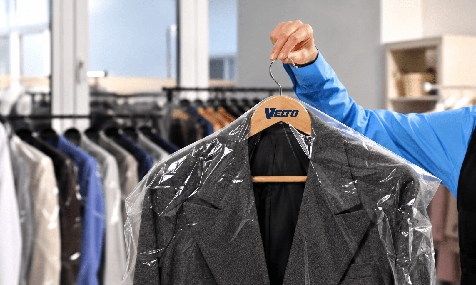 A hand holding up a grey suit jacket in a clear garment cover on a wooden hanger, with covered garments on rails behind.